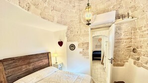 Un dormitorio con pared de piedra, cabecera de madera, cama con edredón blanco, lámpara de noche y un banco.