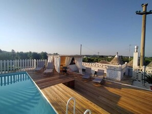 Un espace piscine aménagé sur le toit, avec une terrasse en bois, des chaises longues et un coin salon abrité.