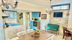 Un comedor con una mesa de madera y sillas blancas, un banco verde a cuadros, y una cocina con armarios azules y una pared de piedra.