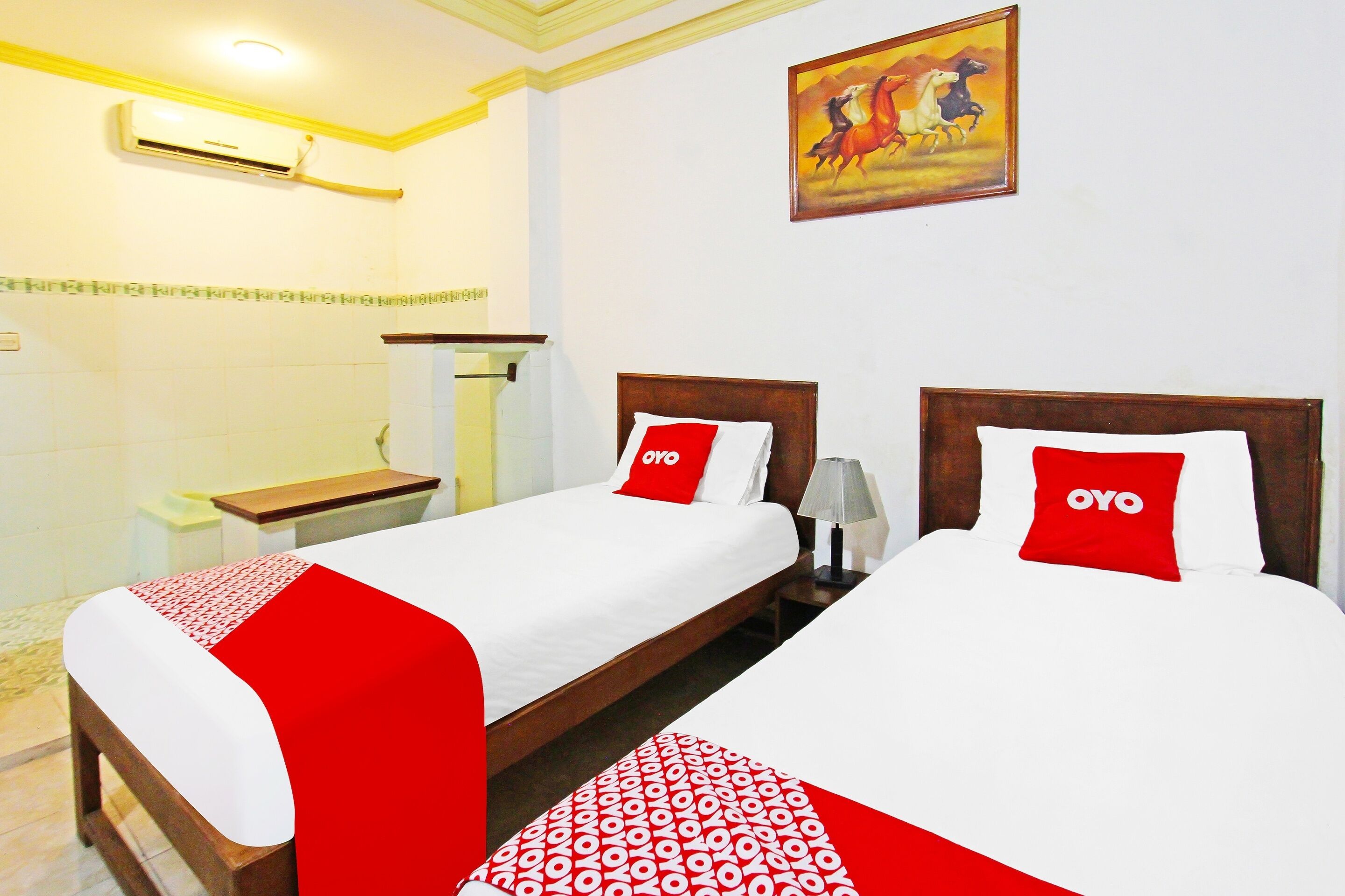 A hotel room with two beds, each with a red and white bedspread and pillows, a wooden headboard, a bedside table, and a wall-mounted air conditioner.