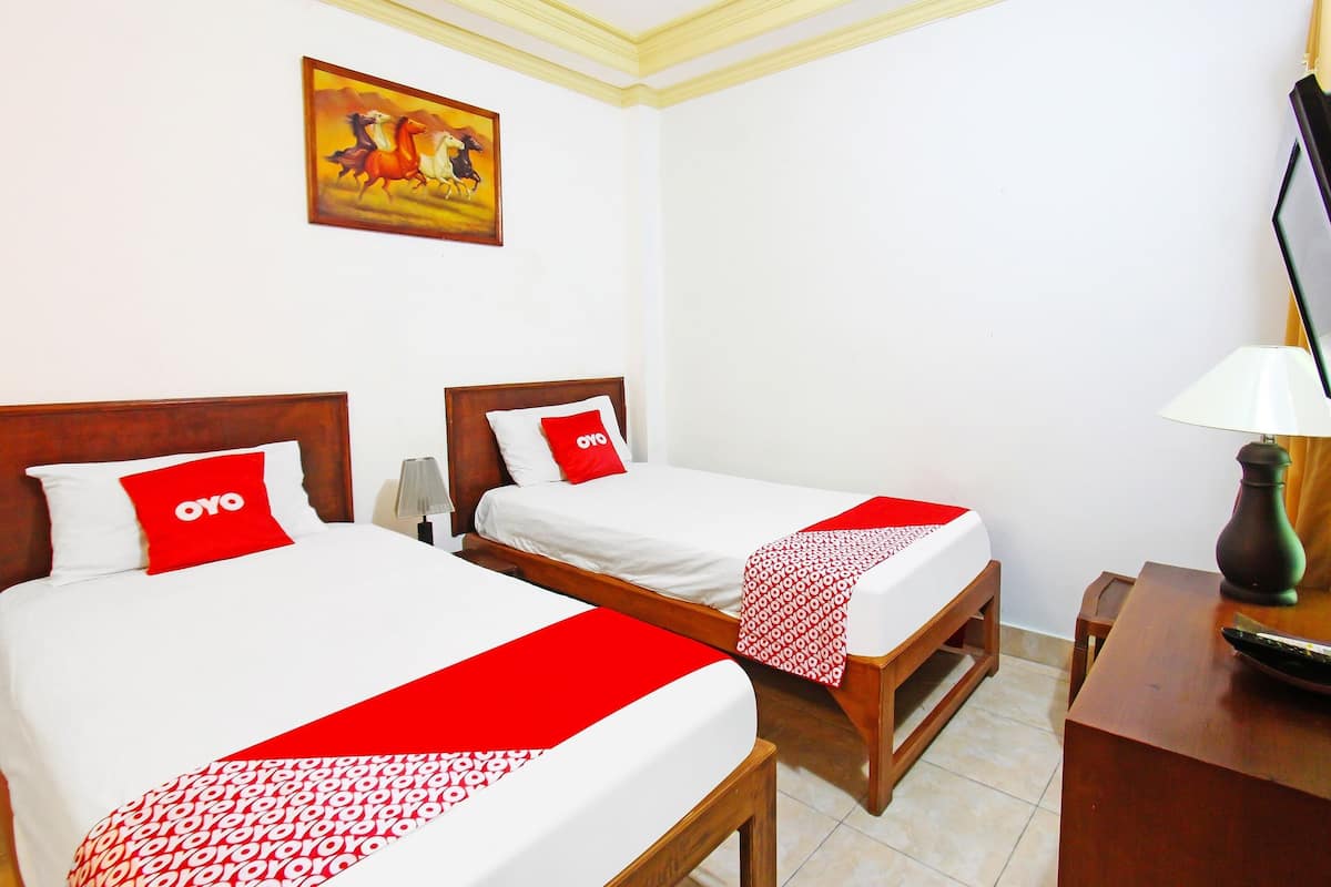 A hotel room with two beds, each with a red and white bedspread and a pillow with the hotel's logo. There is a painting on the wall, a lamp on a wooden table, and a wooden nightstand.