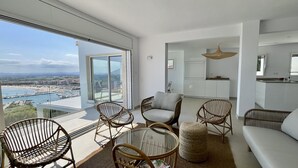 Una sala de estar moderna con una mesa de vidrio, sillas de mimbre y un sofá. Cuenta con un amplio ventanal corredero que da a un balcón con vistas a la ciudad.
