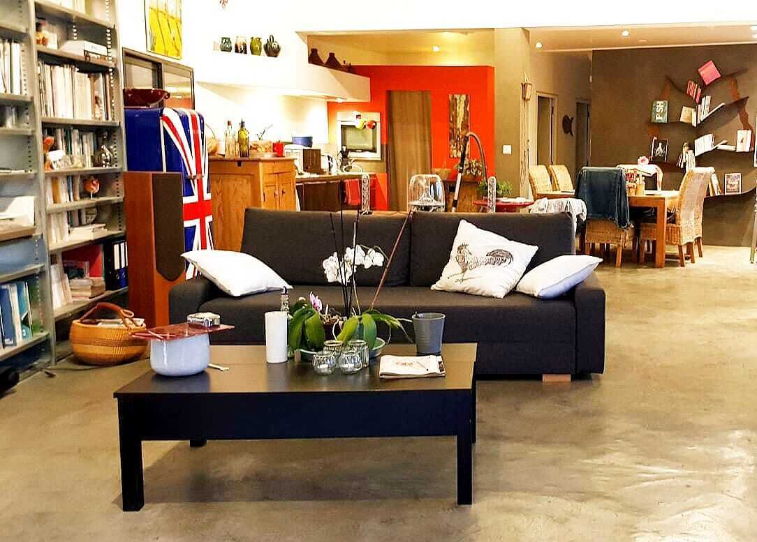 Un salon avec un canapé foncé, une table basse agrémentée d’objets décoratifs, une bibliothèque et, au fond, un coin cuisine.