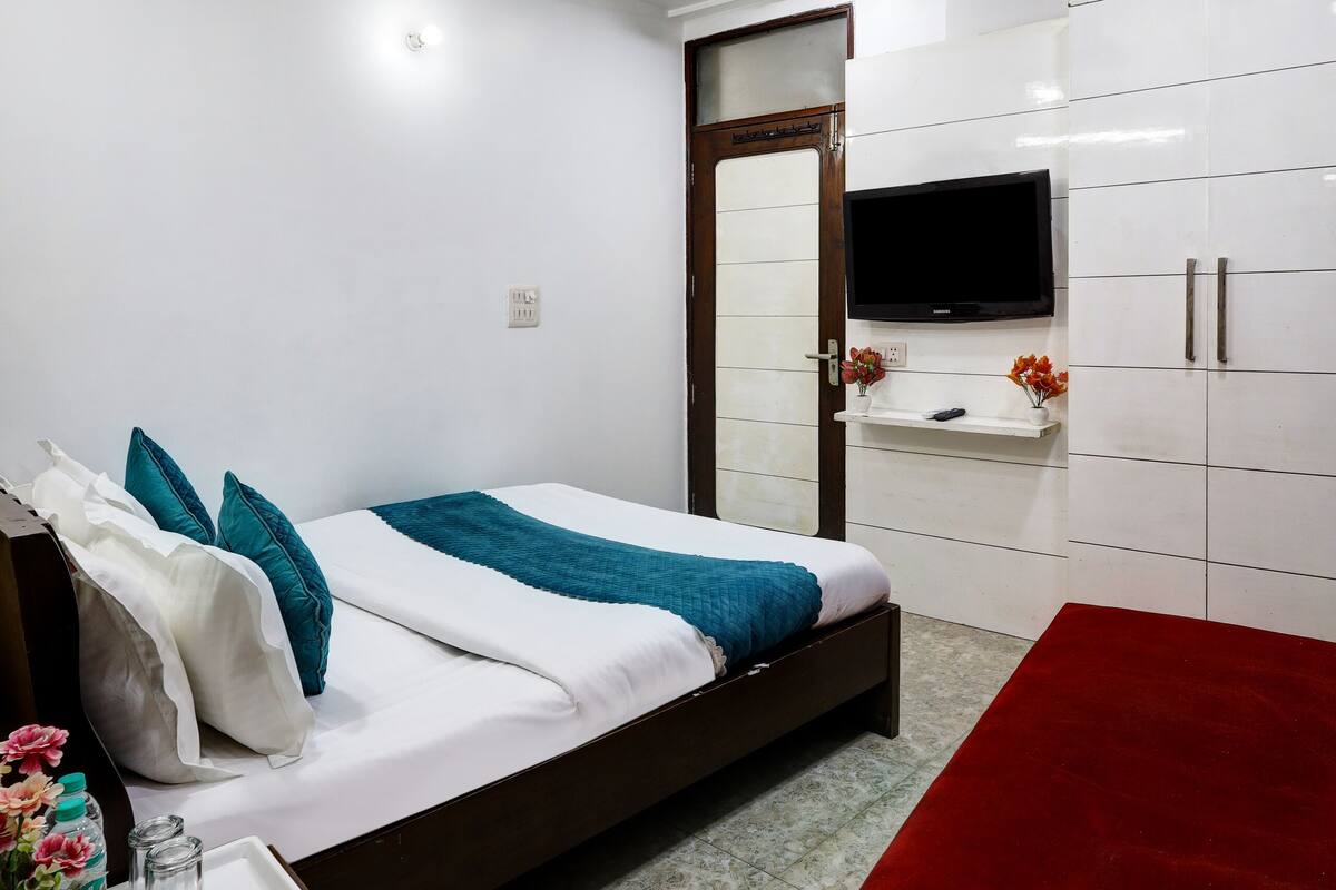 A hotel room with a bed, a red ottoman, a TV mounted on the wall, and a ceiling fan.