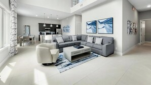 A spacious living area with a grey sectional sofa, a white armchair, and a coffee table. A kitchen is visible in the background with modern appliances.