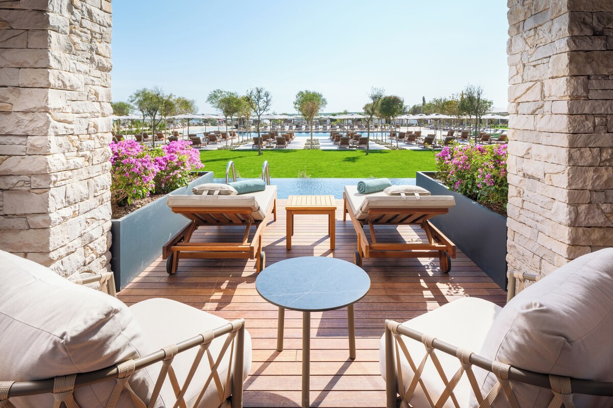 A rooftop terrace with lounge chairs, a small table, and a view of a pool and garden.