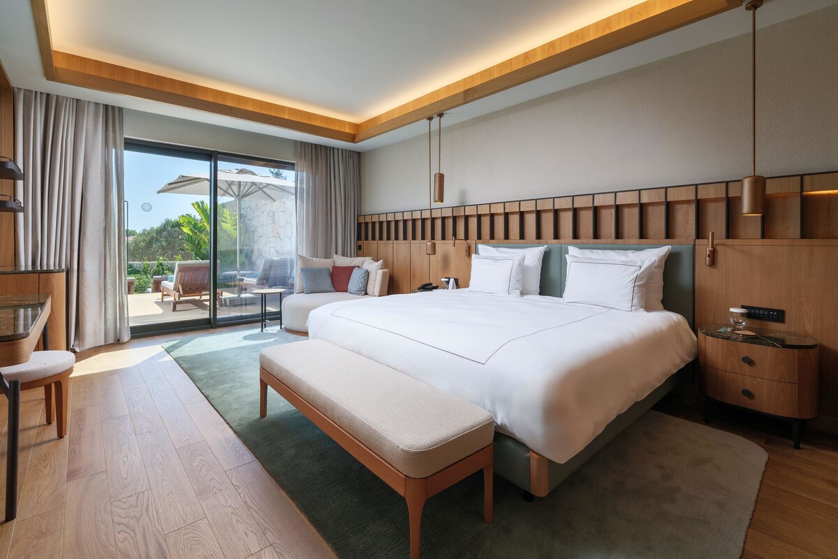 A modern hotel room with a large bed, a desk, a bench, and a view of the outdoors.
