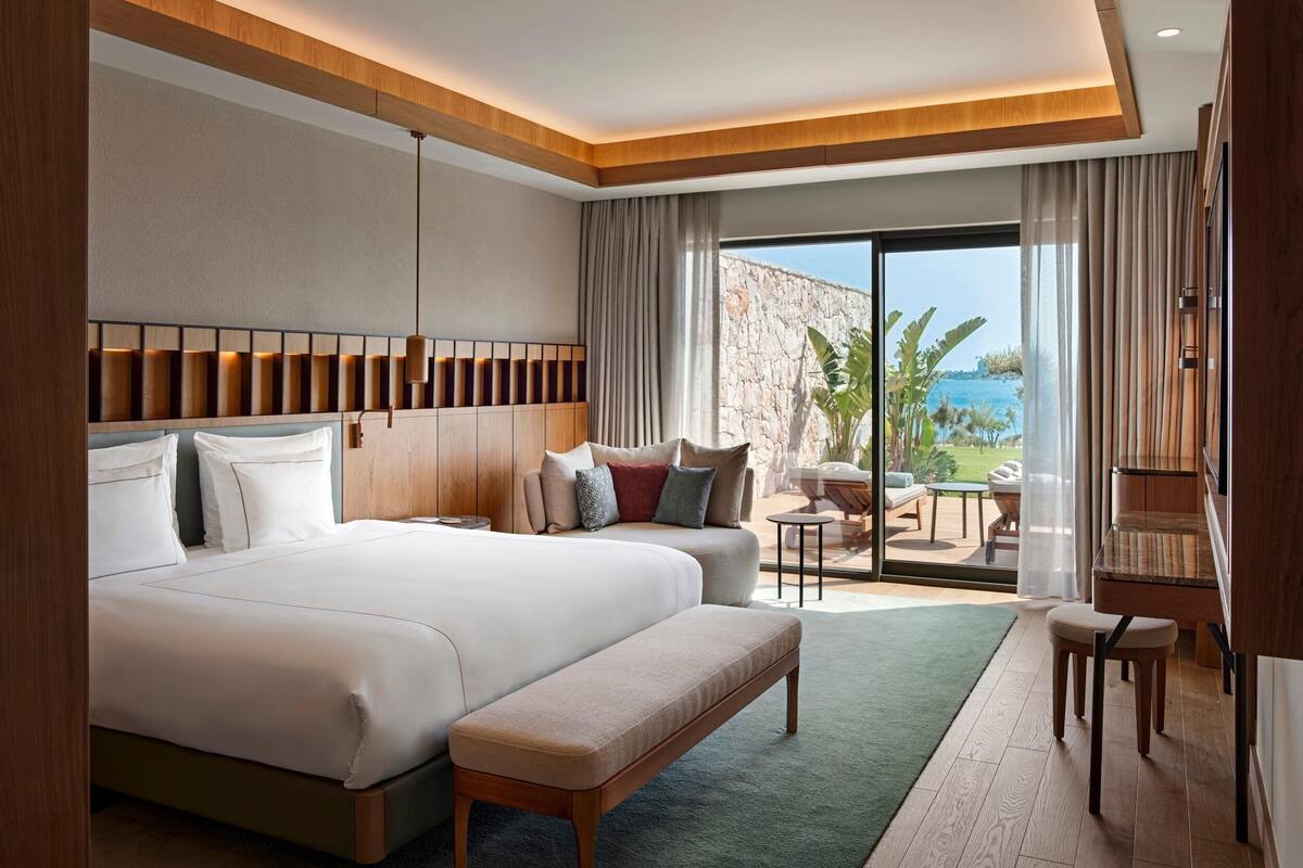 A hotel room with a large bed, a sofa, a bench, a small table, and a view of the sea.