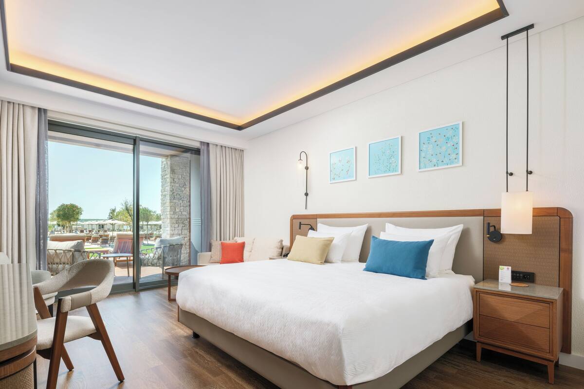 A hotel room with a large bed, a desk, a chair, and a view of the outdoors.