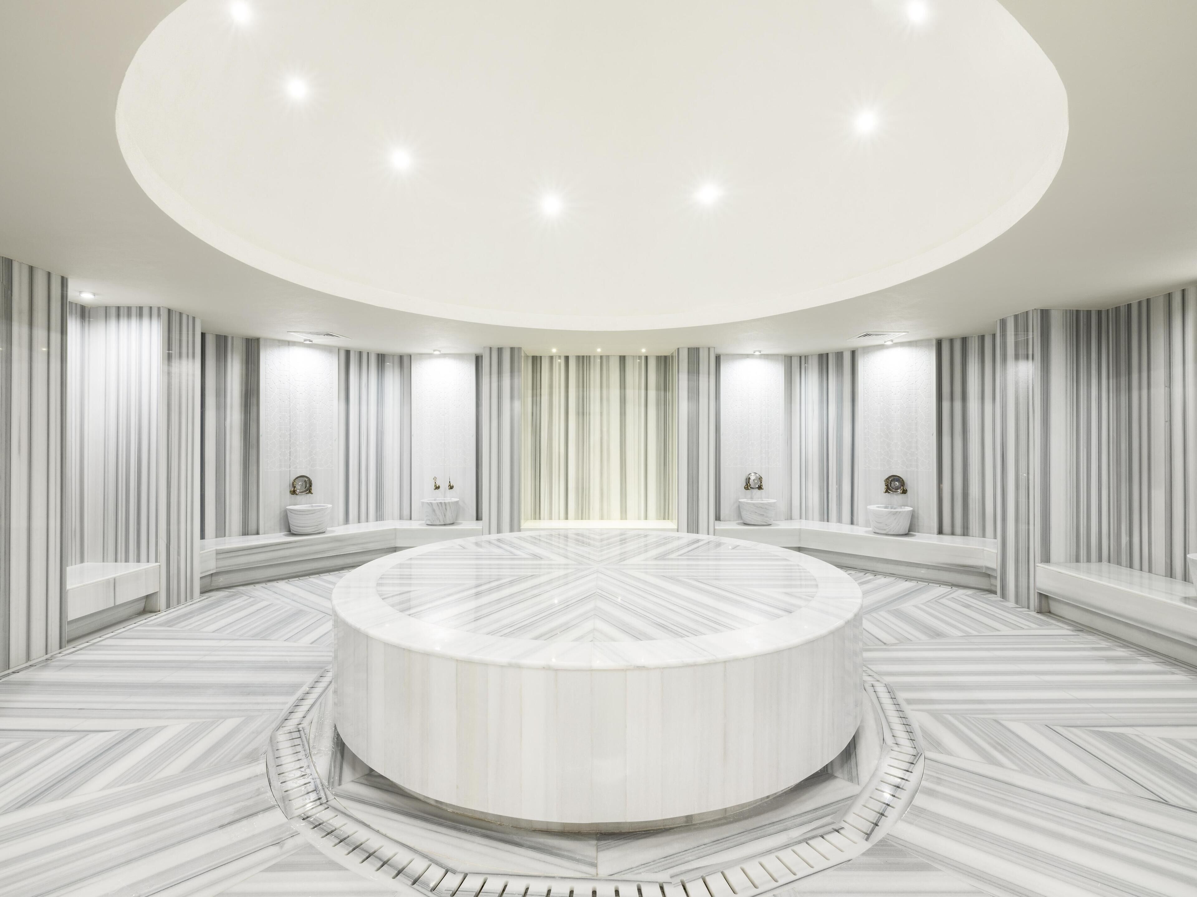 Un área de recepción circular con suelo de mármol, paredes blancas con franjas verticales y una mesa redonda central.