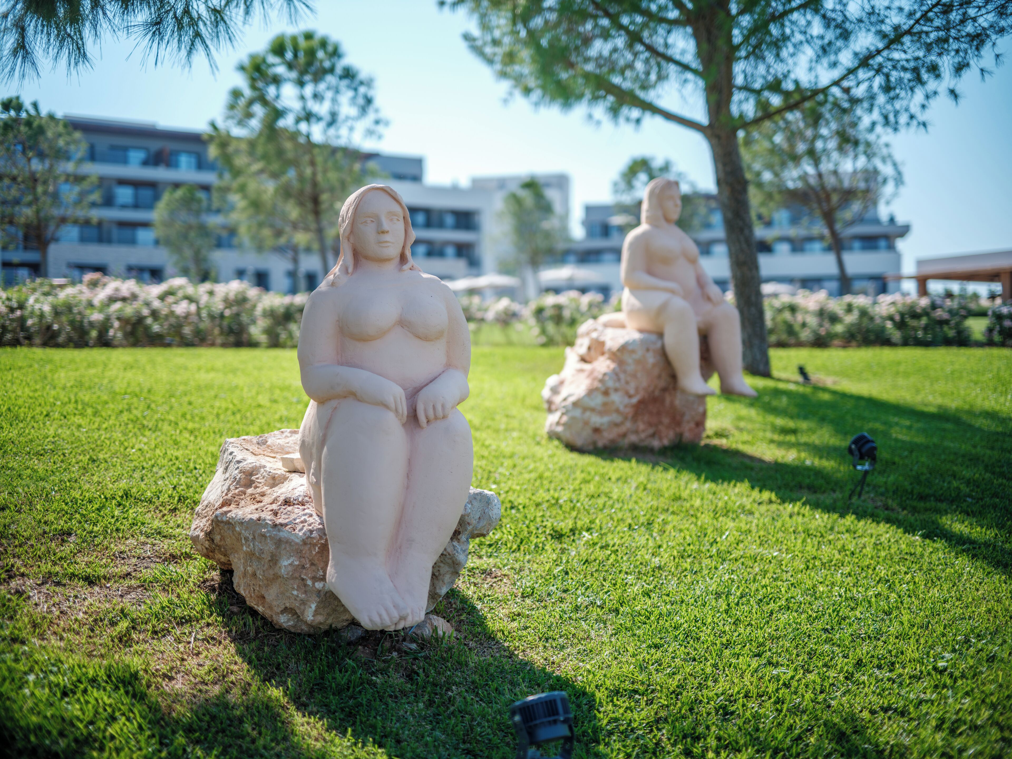 Two nude statues on rocks in a grassy area with buildings in the background.