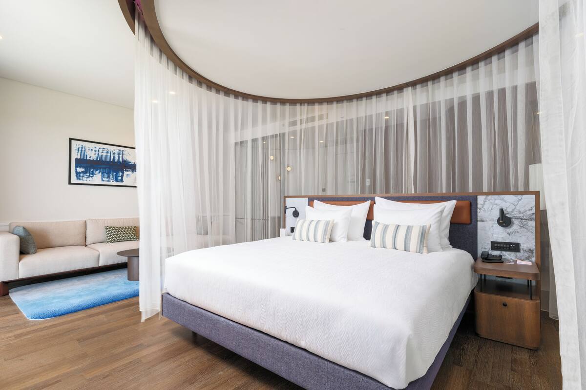 A modern hotel room with a large bed, a bedside table with a phone and a lamp, and a sofa in the corner.