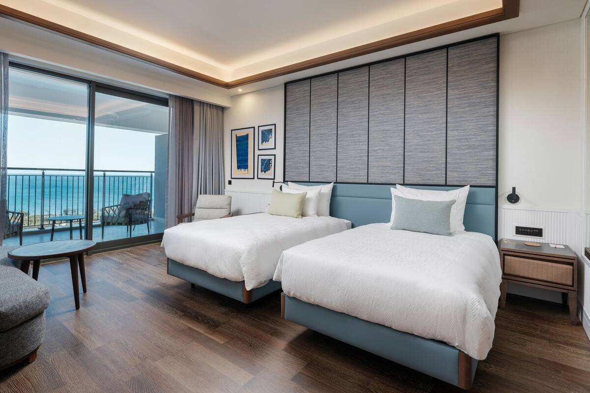 A hotel room with two beds, a balcony with a view, a small table, and a chair.