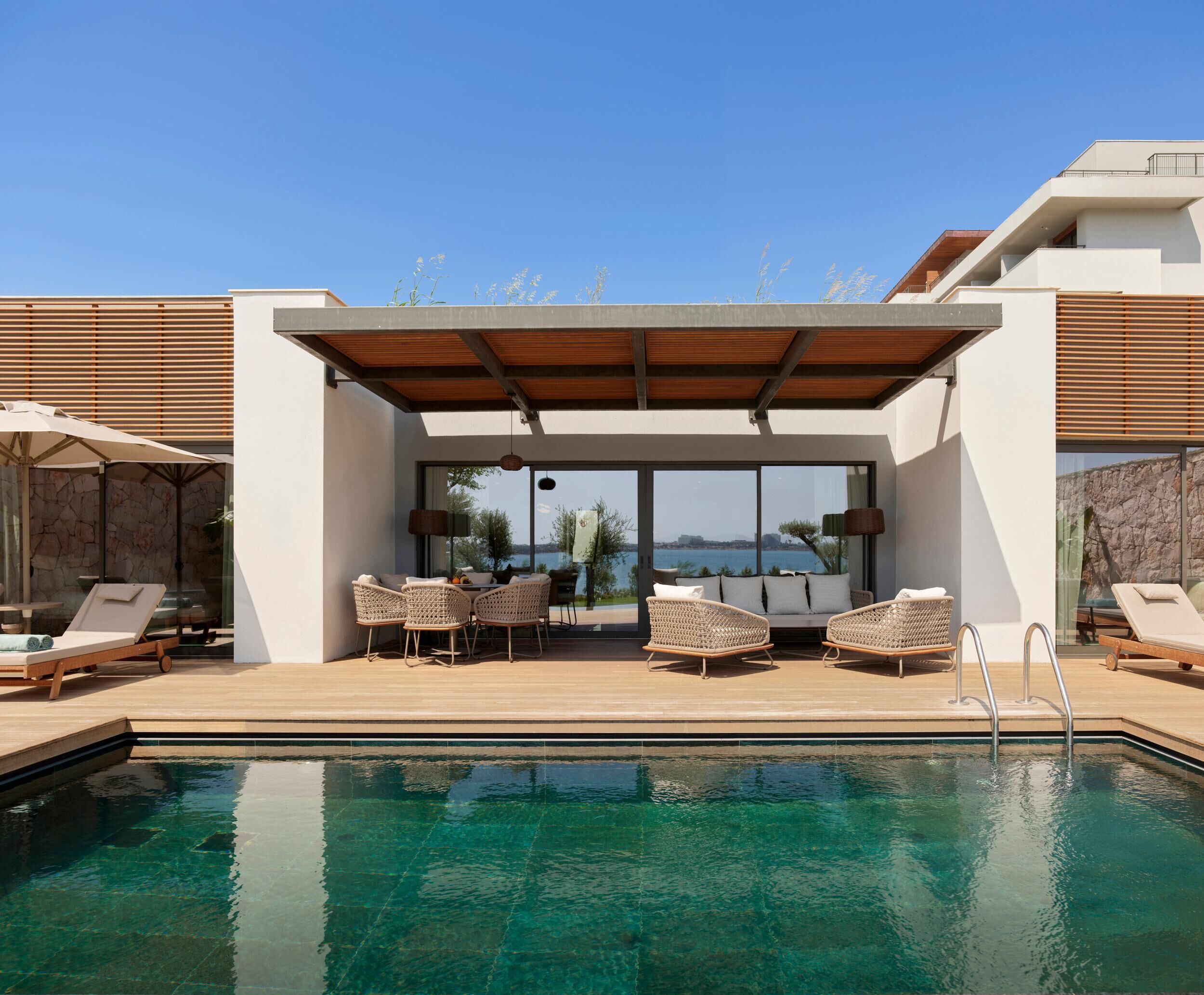 Un área de piscina moderna al aire libre con zona de estar techada, sillones de descanso y vistas al mar.