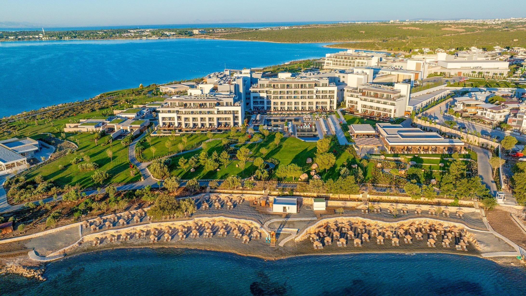 An aerial view of a coastal hotel with multiple buildings, a landscaped garden, and a beachfront with umbrellas.