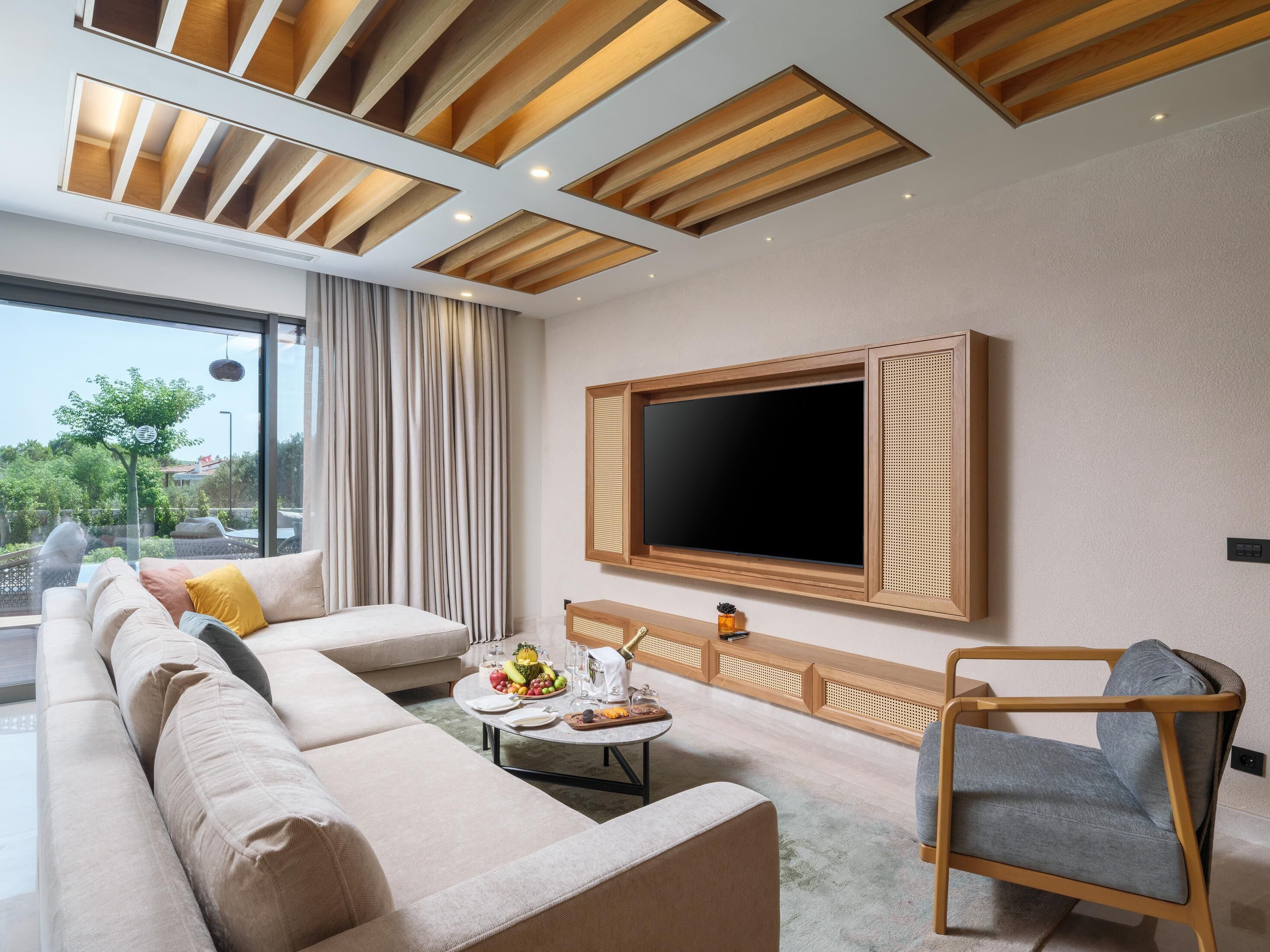 Una sala de estar moderna con un sofá modular grande, una mesa de centro redonda con frutas, una pared de acento de madera y un televisor grande montado en la pared.