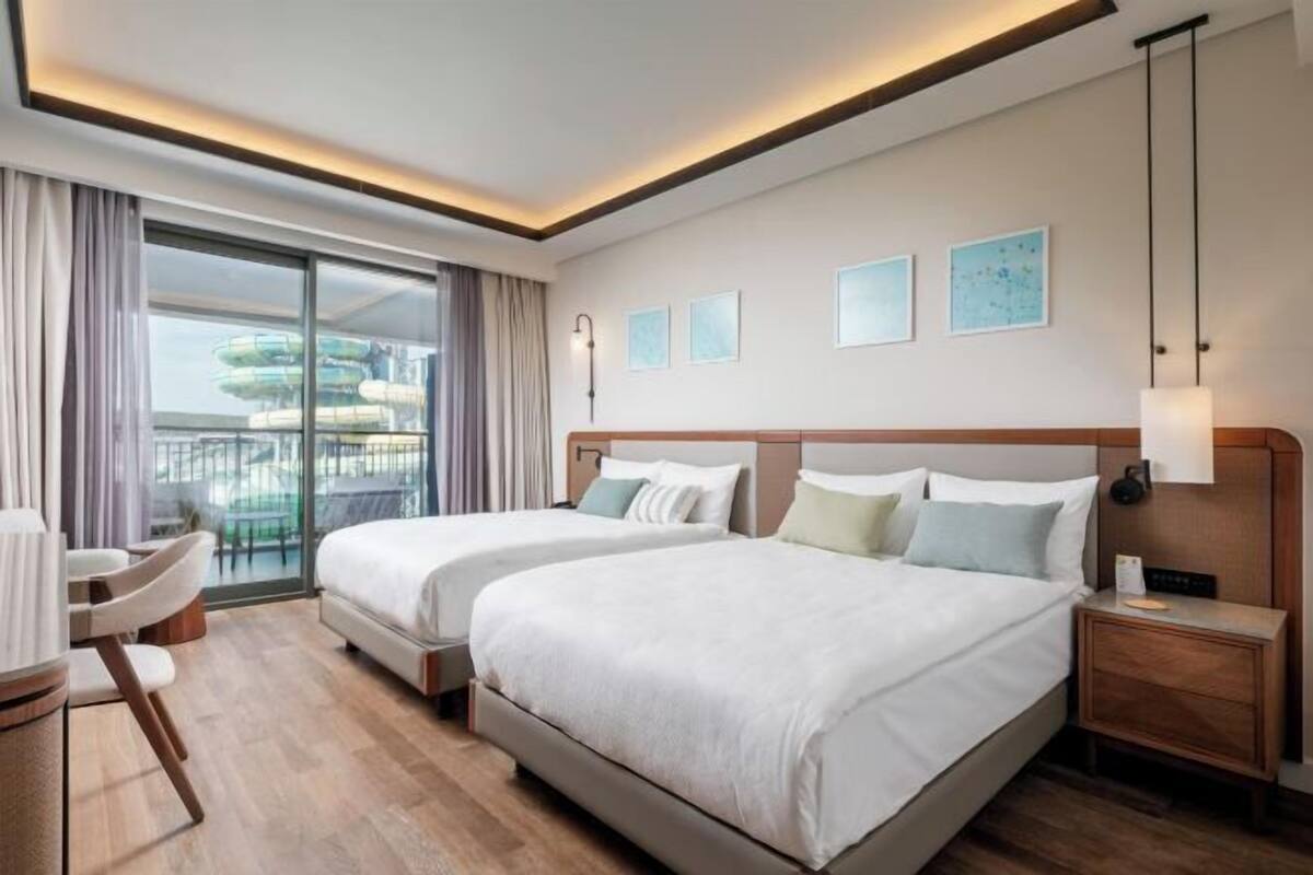 A hotel room with two beds, a desk, a chair, a nightstand, and a view of a water park.