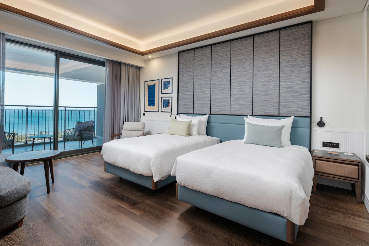 A hotel room with two beds, a balcony with a view, a small table, and a chair.