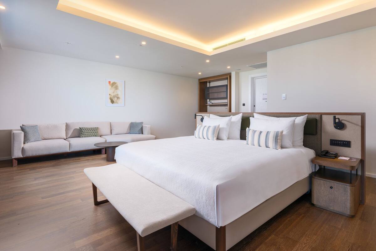 A modern hotel room with a large bed, a bench, a sofa, a bedside table, and a mirror.