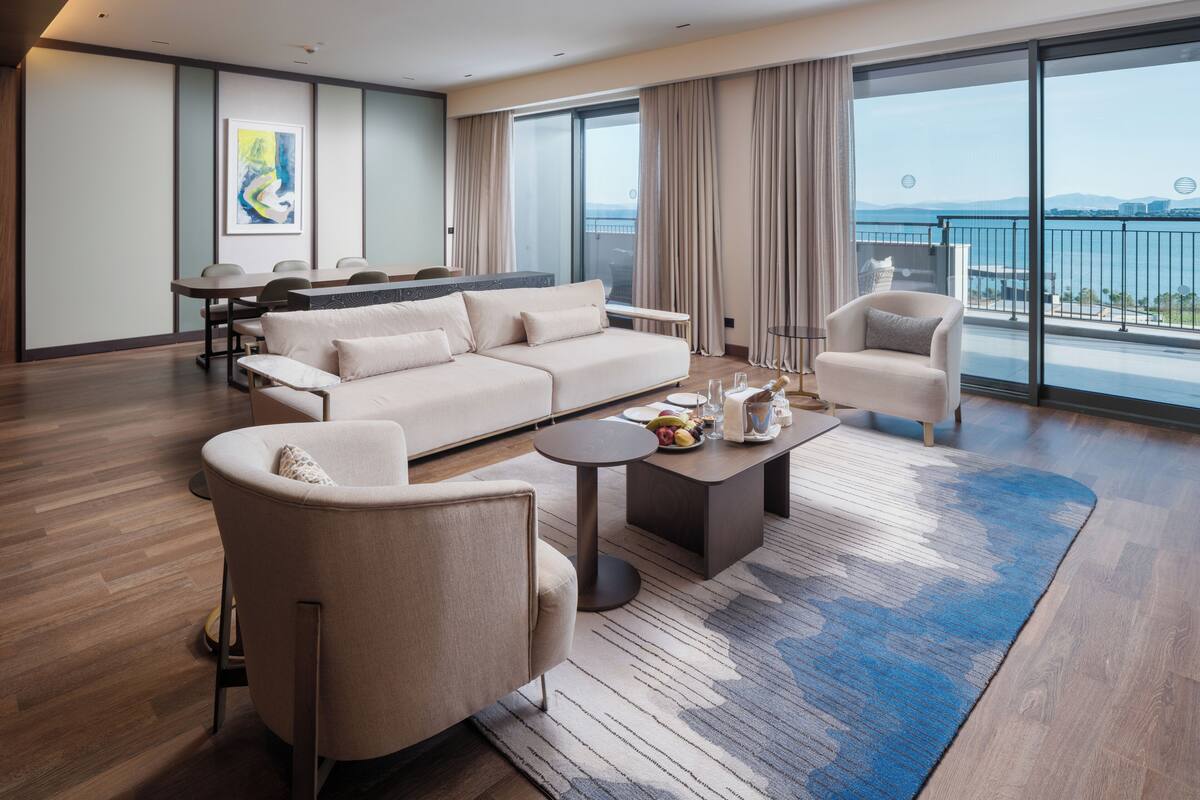 A modern hotel room with a large sofa, armchairs, a dining table, and a balcony with a view of the sea.