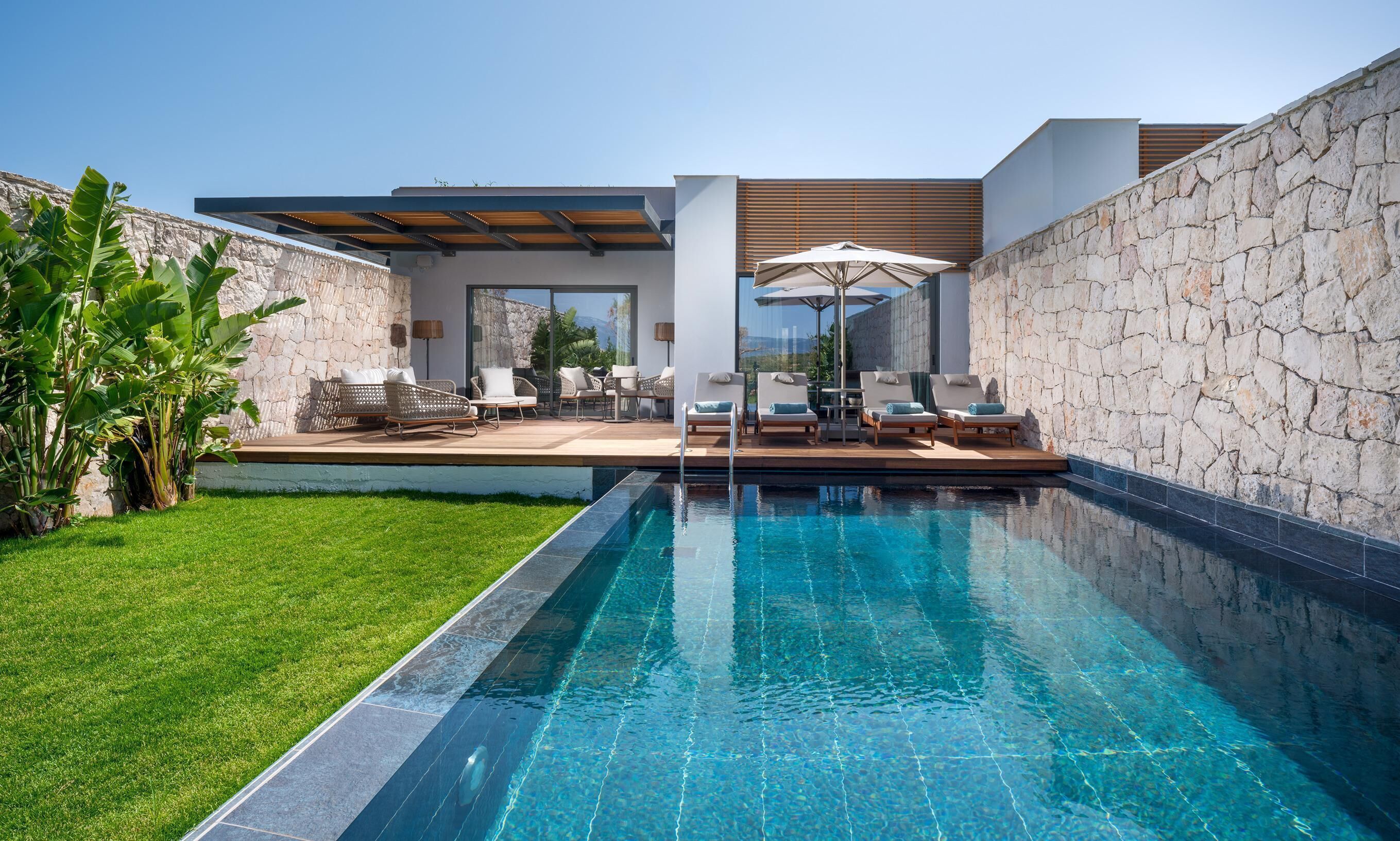 Zona de piscina exterior moderna con un muro de piedra, una terraza de madera y sillas de descanso.