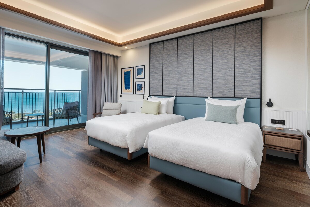 A hotel room with two beds, a balcony with a view, a small table, and a chair.