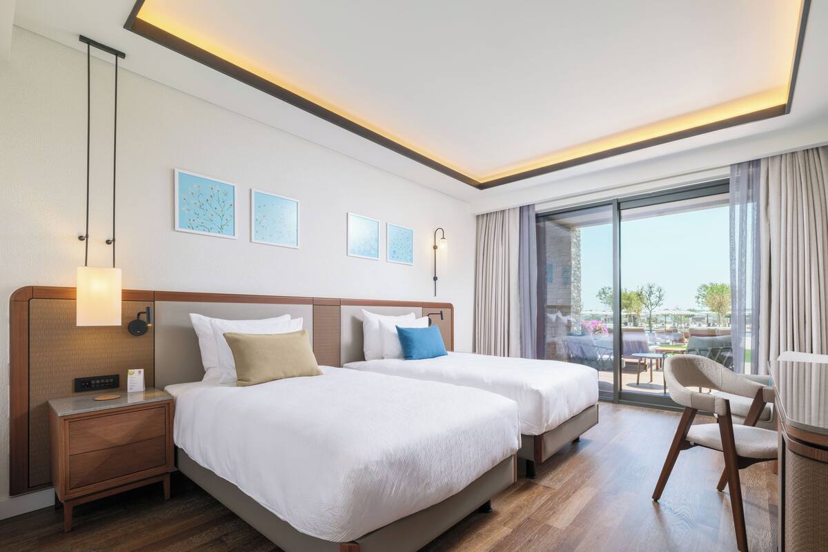 A hotel room with two beds, a nightstand, a desk, and a chair. There is a large window with a view of the outside.