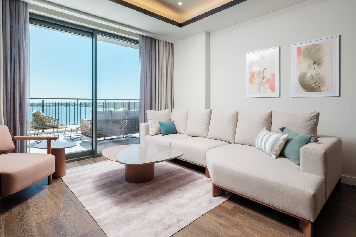 A modern living room with a large sofa, a coffee table, and a view of the sea through a balcony.