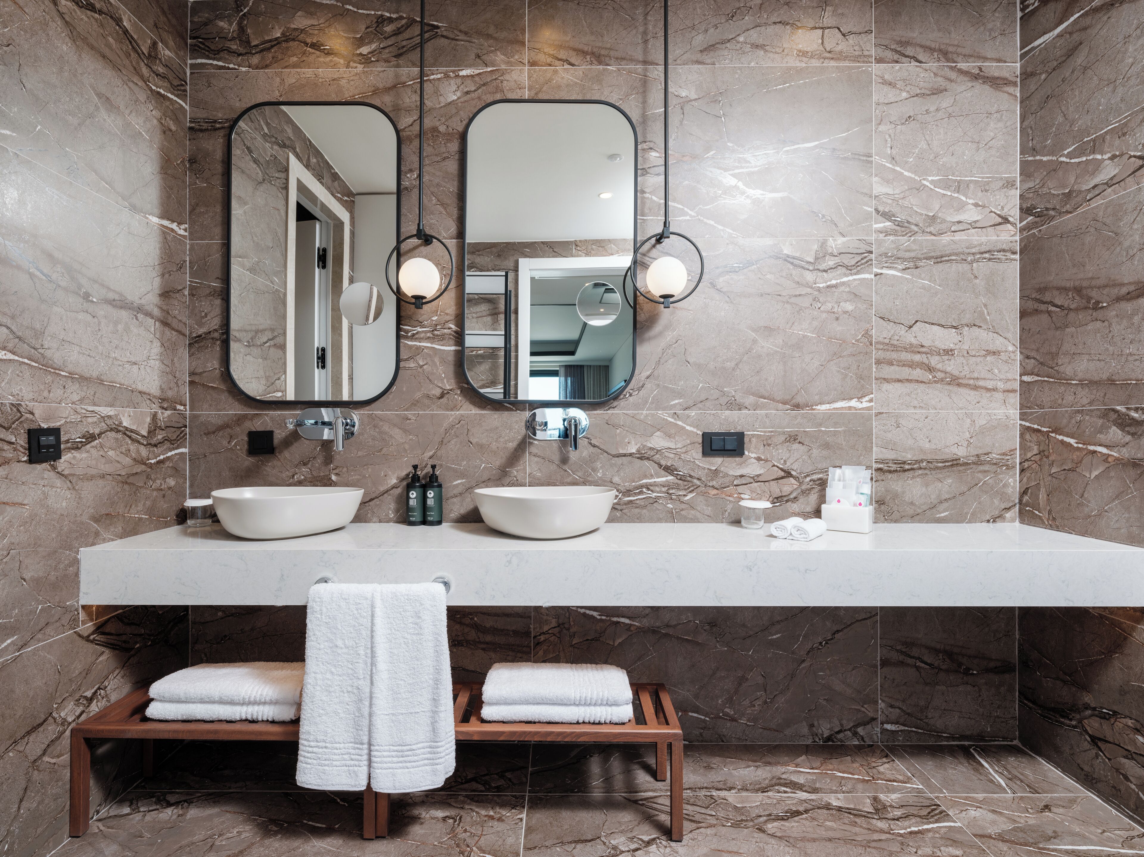 A modern bathroom with two sinks, a large mirror, and a wooden bench with towels.