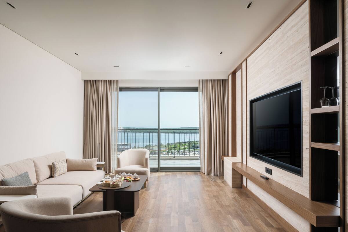 A modern living room with a large TV, a sofa, a coffee table, and a view of the sea through large windows.