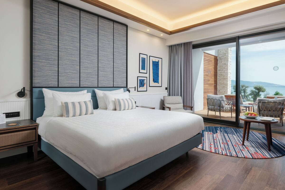A modern hotel room with a large bed, a view of the sea, and a balcony with outdoor seating.