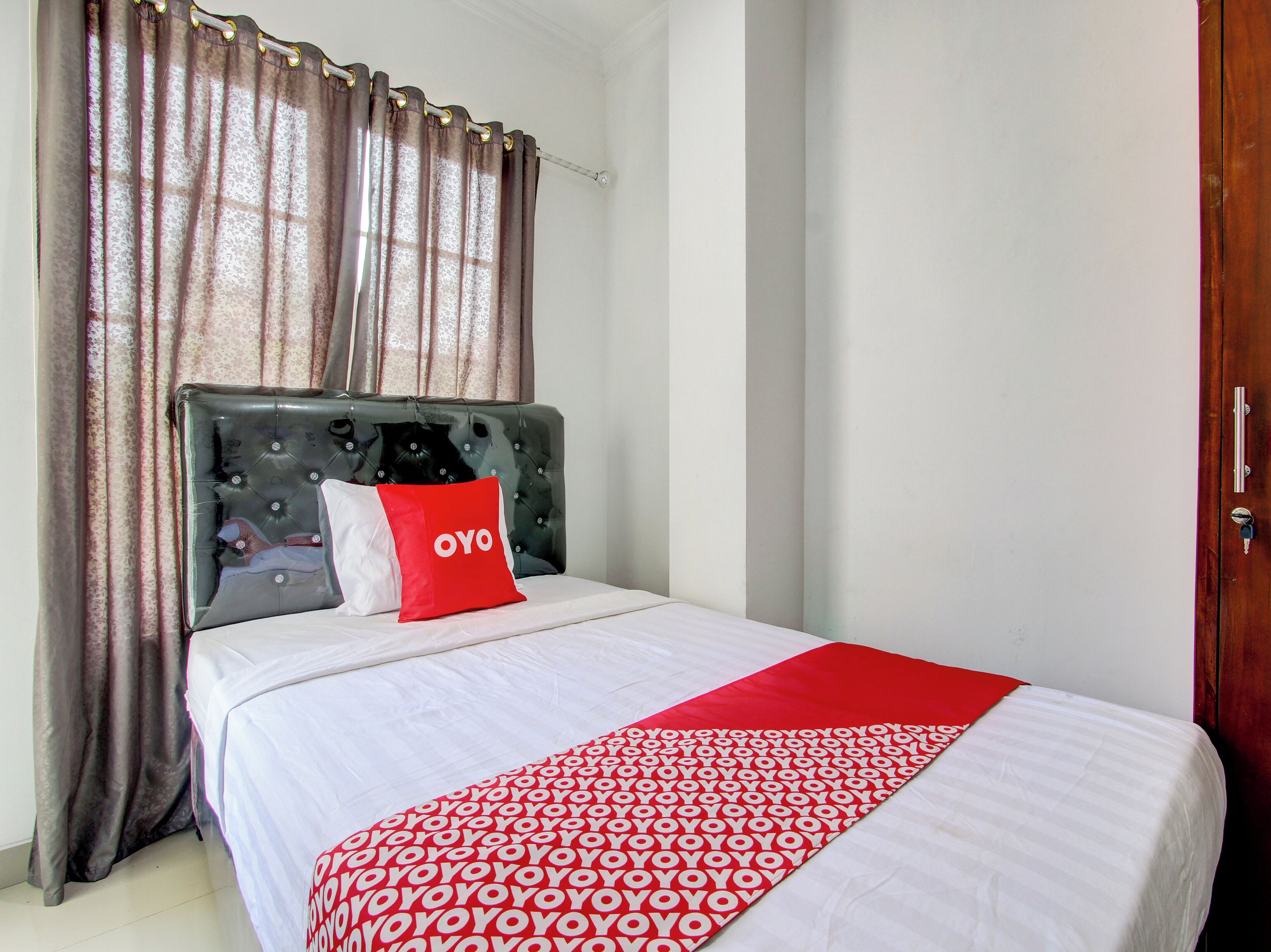 A neatly made bed with a white and red patterned bedspread, a red pillow with "OYO" text, and a tufted headboard.
