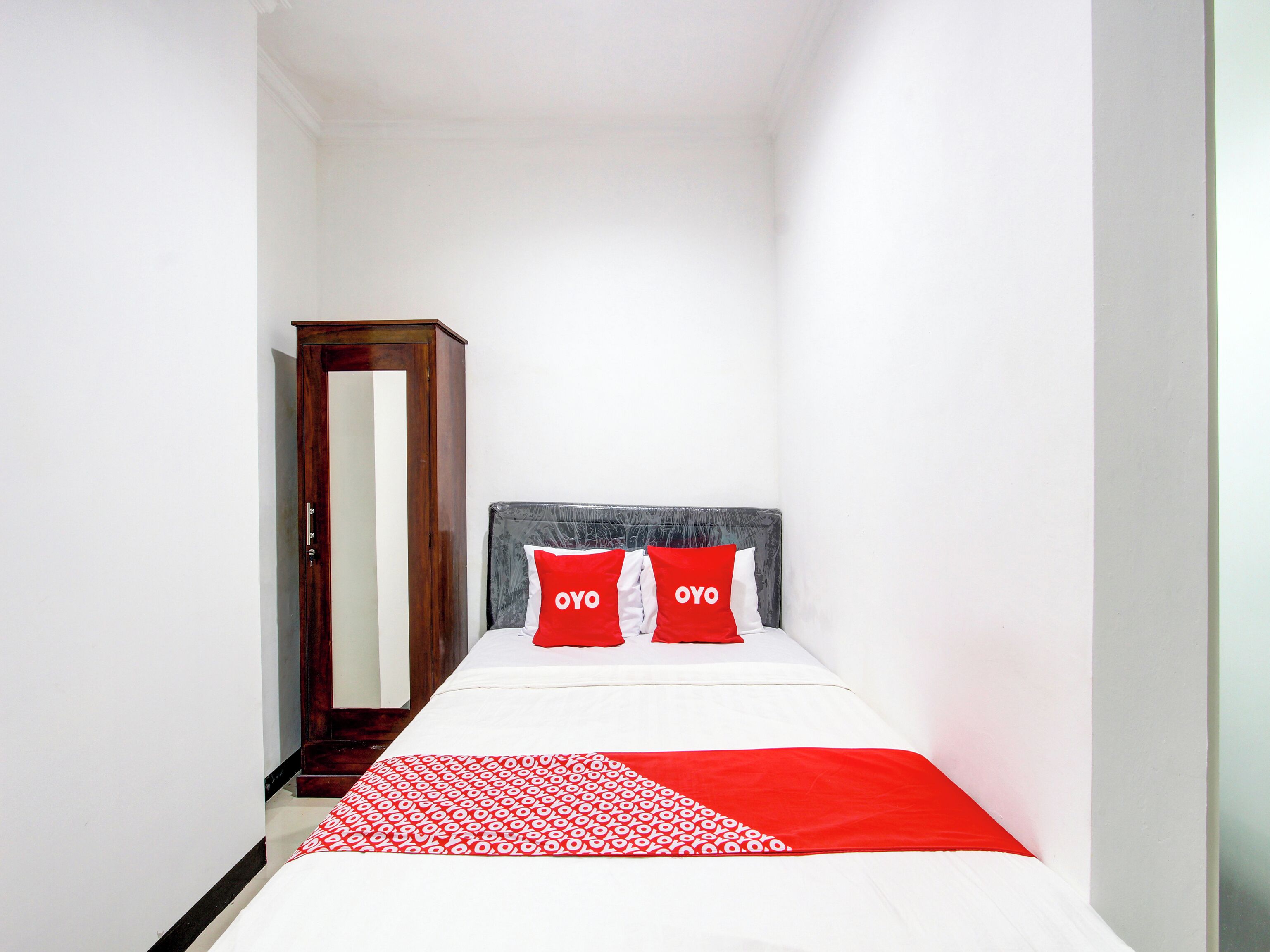 A narrow hotel room with a bed, two red pillows with "OYO" text, a wooden wardrobe, and a white wall.