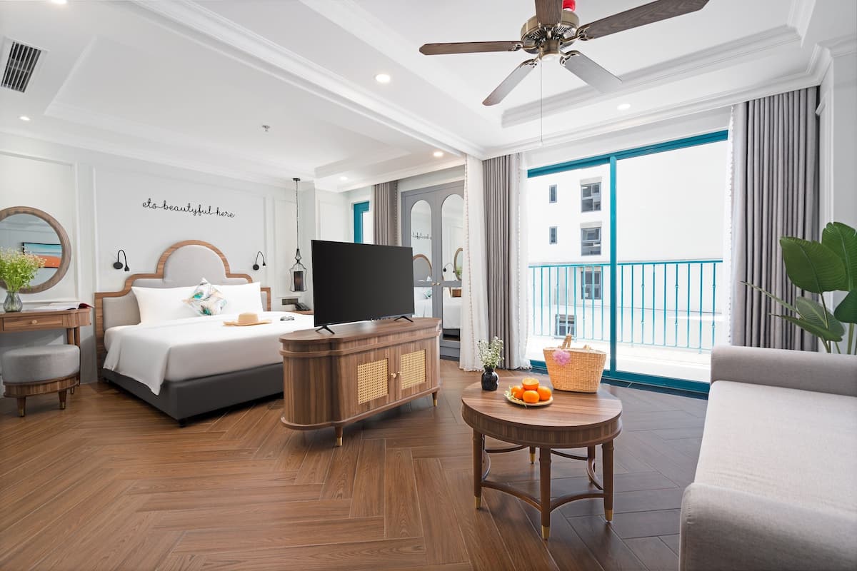 Habitación de hotel con cama, televisor, cómoda de madera, mesa redonda y balcón con vistas.