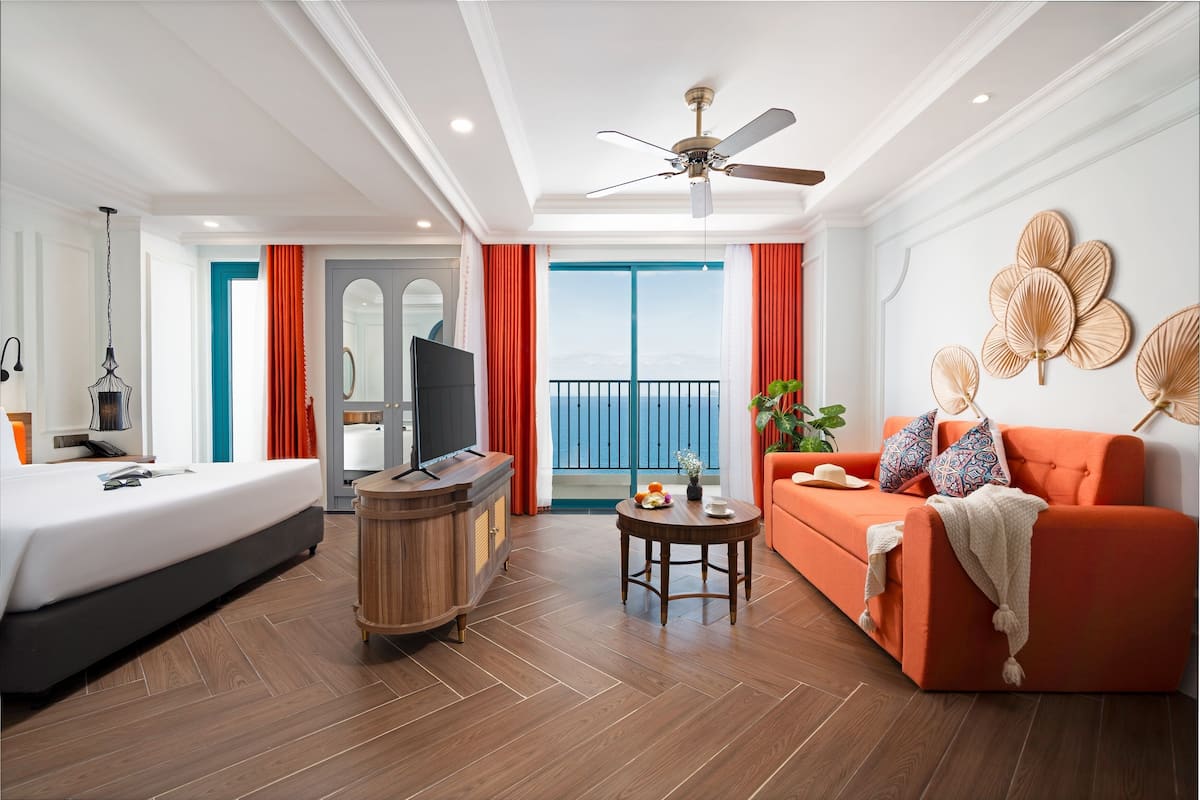 Habitación de hotel con cama, sofá, televisión, mesita de madera y balcón con vistas.
