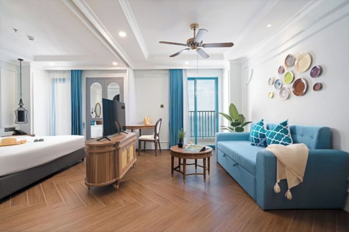A modern hotel room with a bed, a blue sofa, a wooden cabinet, a small table, and a ceiling fan.