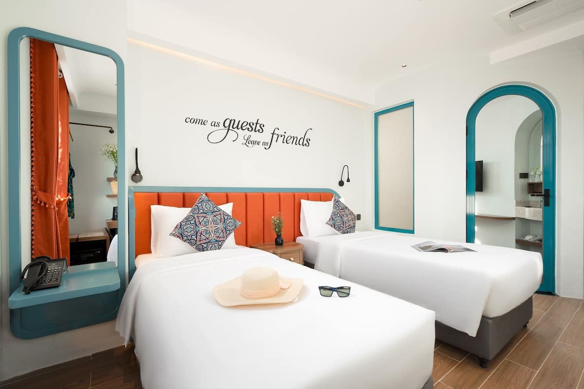 Habitación de hotel con dos camas, un espejo, un cartel en la pared, un teléfono, un sombrero y gafas de sol sobre la cama.