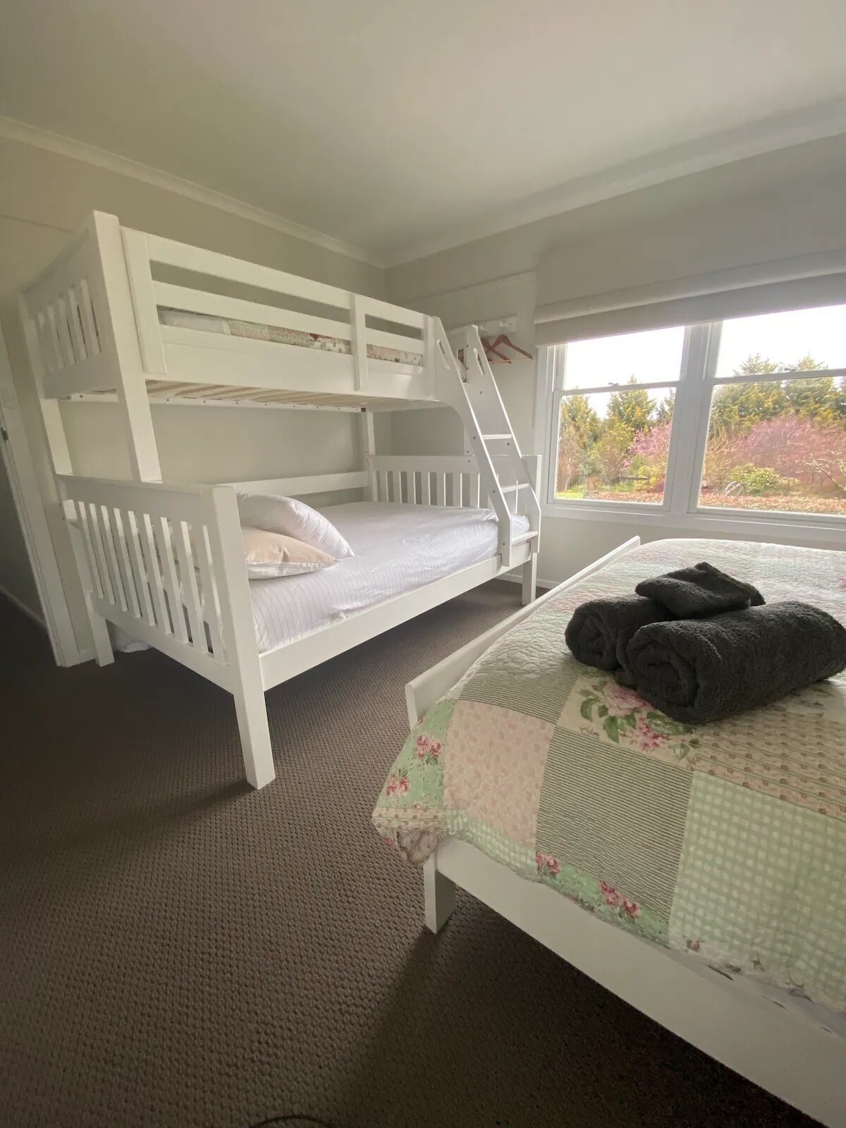 A room with a bunk bed, a window with a view of trees, and a bed with a floral quilt.