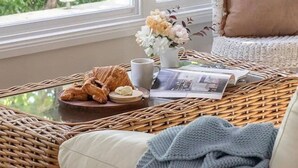 Una habitación acogedora con una silla de mimbre, un cojín, una mesita con un croissant, una taza y una revista.
