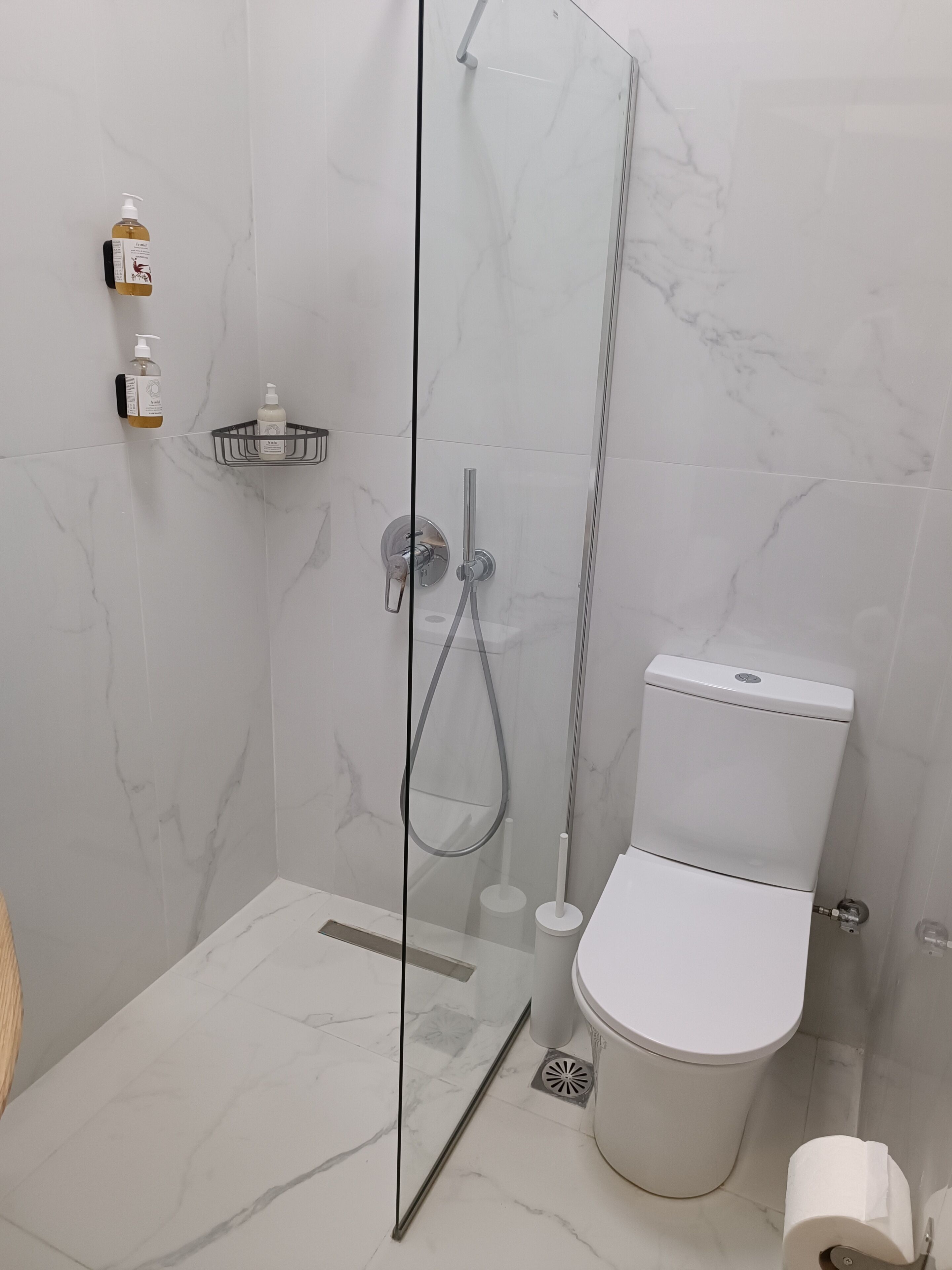 A modern bathroom with a glass-enclosed shower, a toilet, and a small shelf with toiletries.