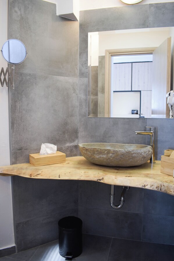 A bathroom with a large round mirror, a stone vessel sink, a wooden box, and a black trash can. - Ulysses Apartments Acropolis (Athens)