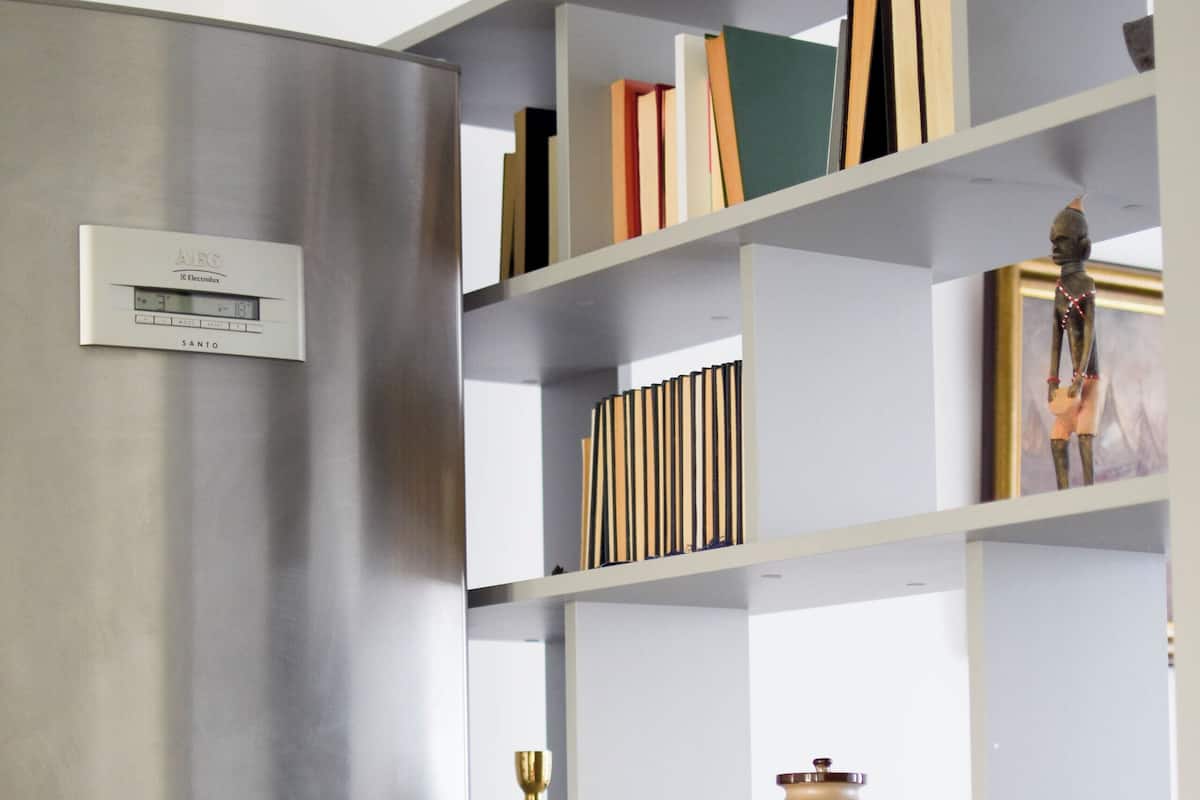 A modern kitchen with a stainless steel refrigerator, a built-in bookshelf with various items, and a coffee maker on the countertop.