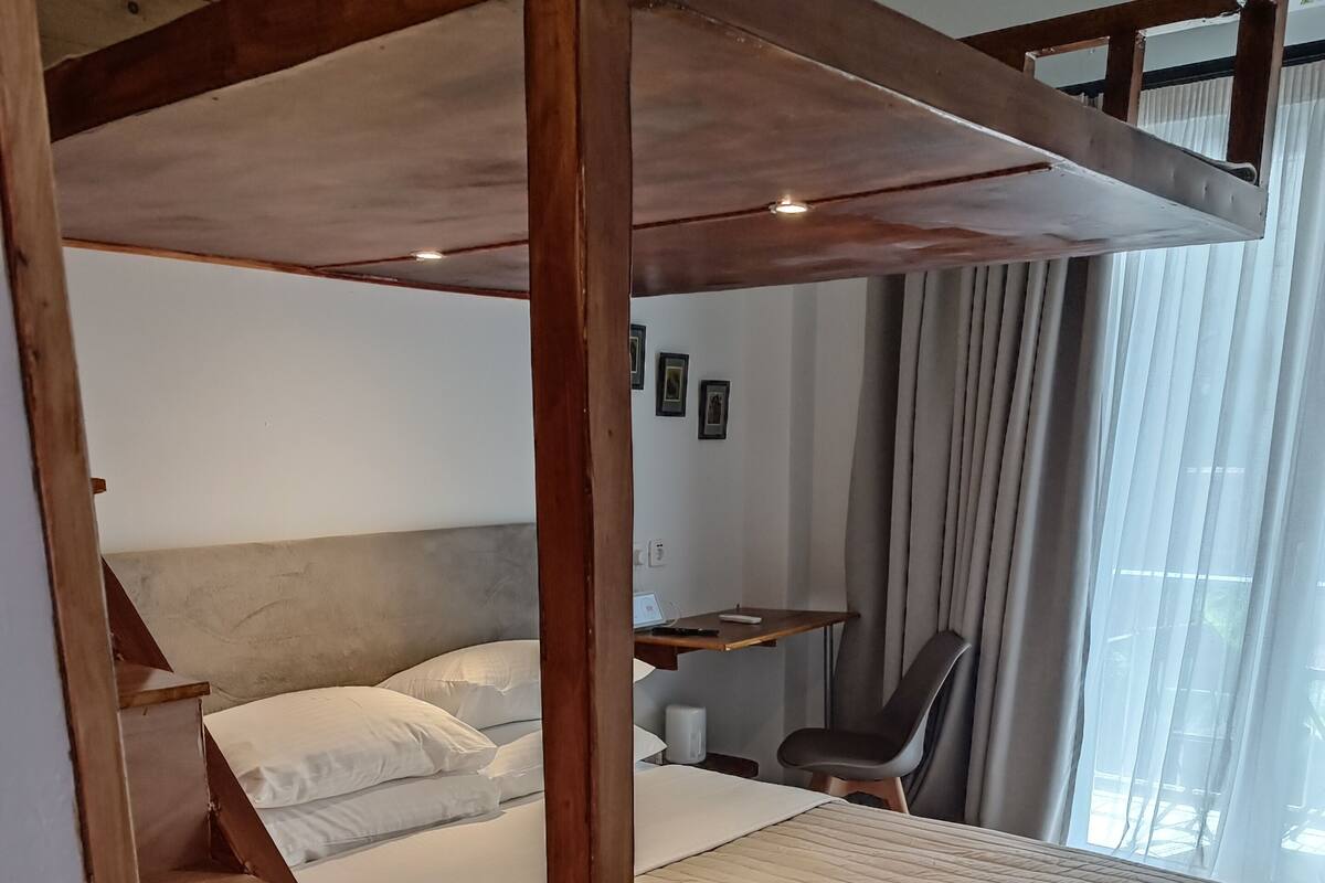 A bedroom with a wooden staircase, a bed with a quilted cover, a desk with a chair, and a window with curtains.