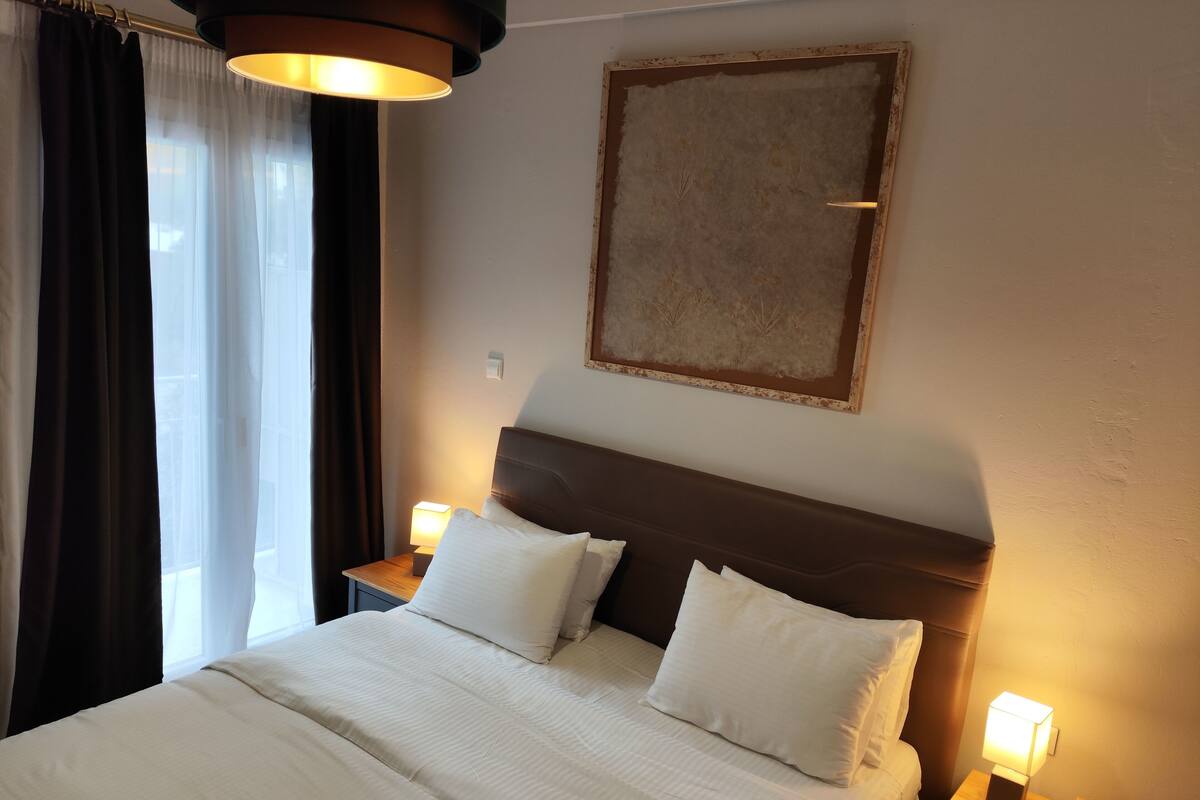 A hotel room with a large bed, two bedside tables with lamps, a painting on the wall, and a window with curtains.