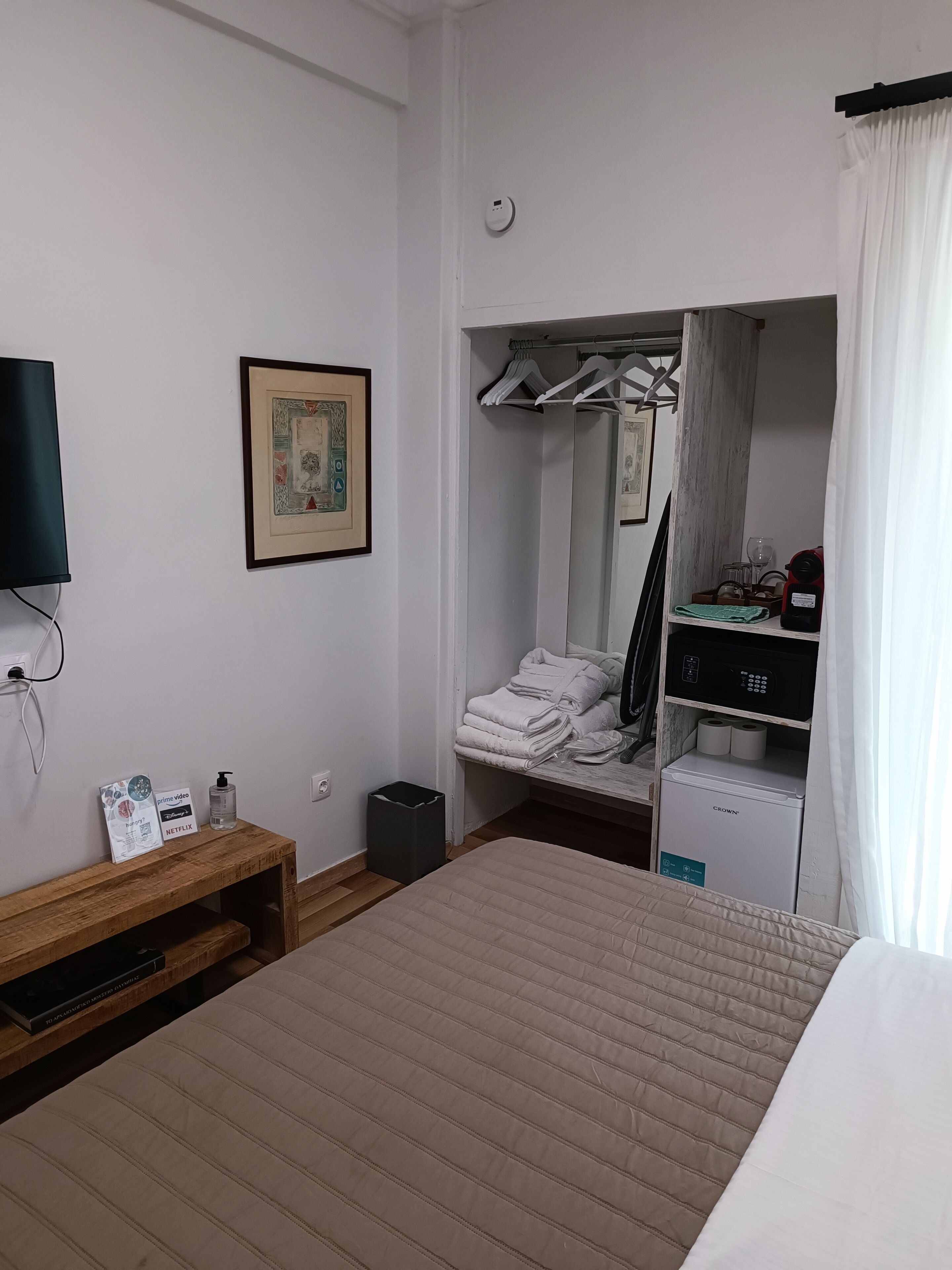 A hotel room with a bed, a wooden nightstand, a framed picture, a mirror, a wardrobe, and a minibar.