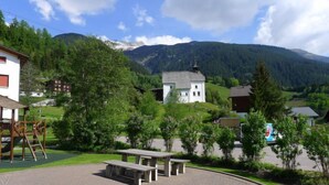 A rural setting with a white building, possibly a church, surrounded by greenery and mountains in the background. - Apt D 016 by Interhome (Disentis)