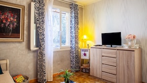 Una habitación con televisión, un armario, una lámpara, un cuadro enmarcado y una ventana con cortinas.