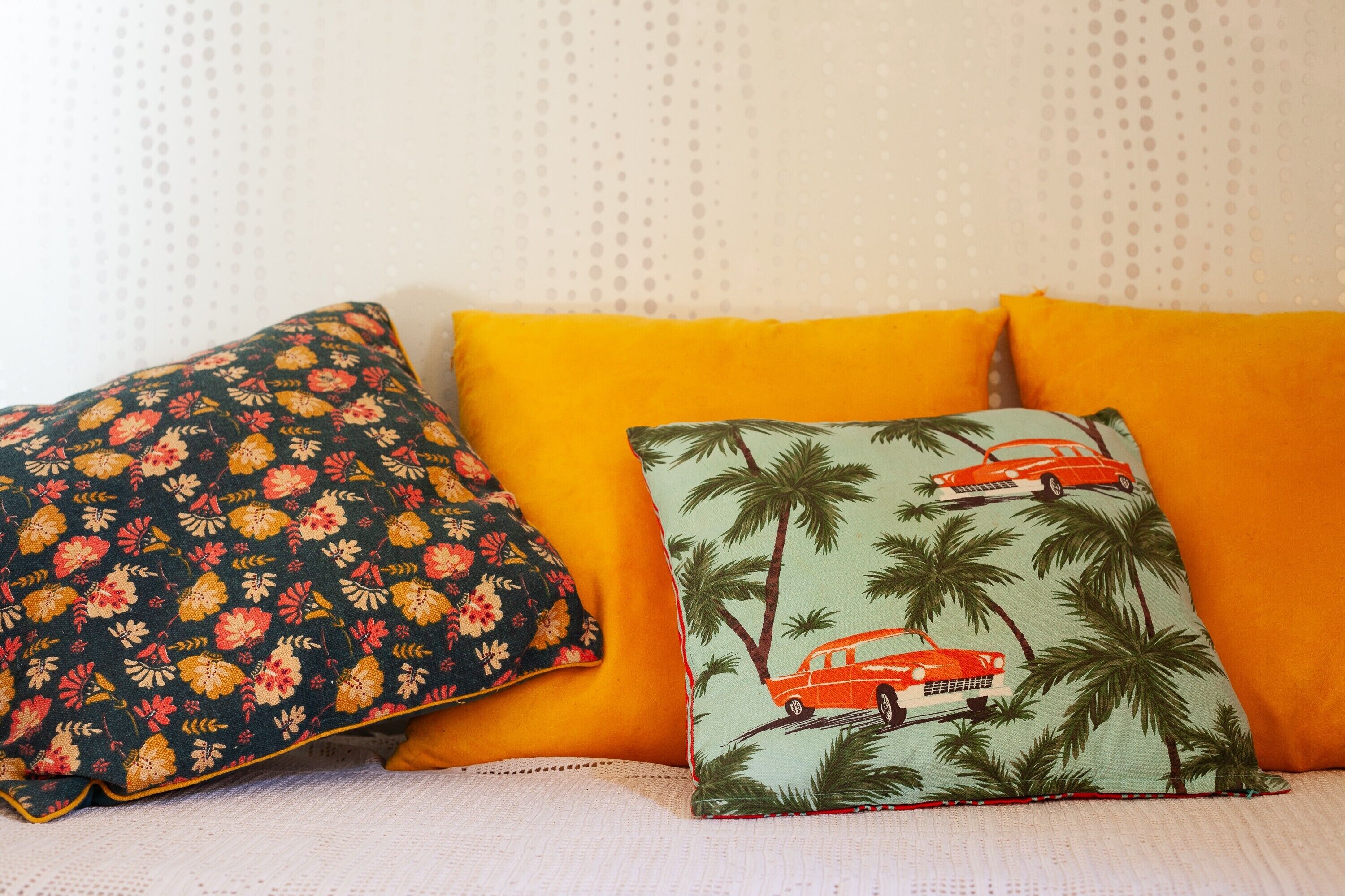 A bed with three pillows, one floral, one with palm trees and a car, and one solid orange.