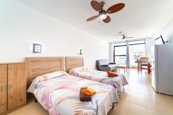 A hotel room with two beds, a ceiling fan, a kitchenette, and a view of the city.
