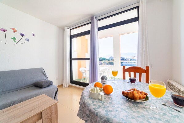 A room with a sofa, a dining table with breakfast items, and a view through a large window.